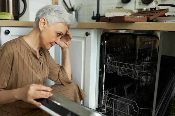 A woman holding her hand on her head while looking disappointed at her dishwasher A woman holding her hand on her head while looking disappointed at her dishwasher
