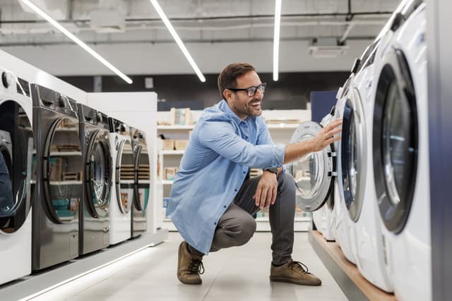 View of a man kneeling down and looking at washing machines in an appliance store View of a man kneeling down and looking at washing machines in an appliance store
