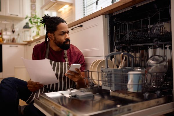 A man in an apron with a paper and phone in hand looking into a dishwasher 