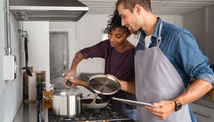 A couple stirs a pot on a stove together and adds salt.