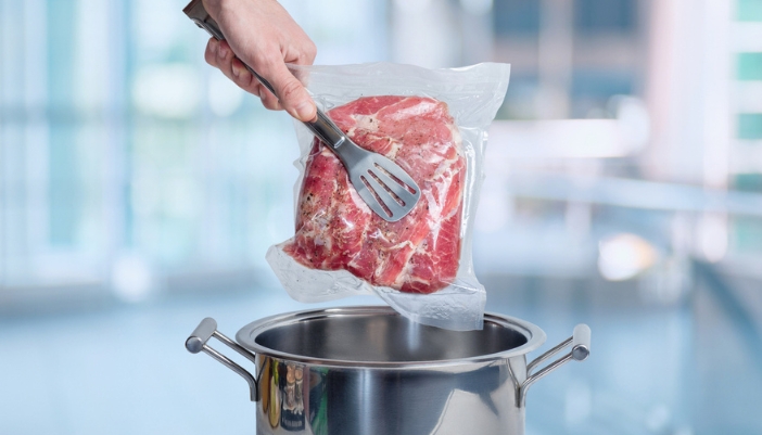 A pair of tongs reach into a pot to grab a vacuum-sealed steak.
