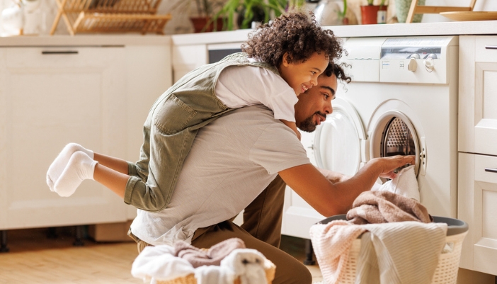 A son rides on his father’s back as they empty a laundry machine. 