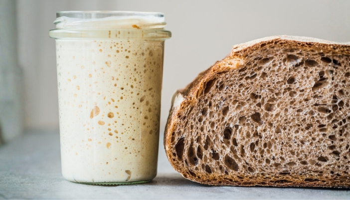 A sourdough starter in a jar sits next to a cut open loaf of sourdough bread.