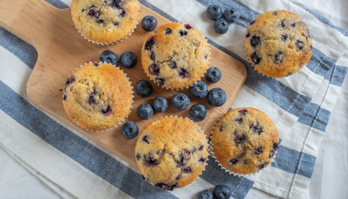 A tray of blueberry muffins rests on a table with blue accents and blueberries around.