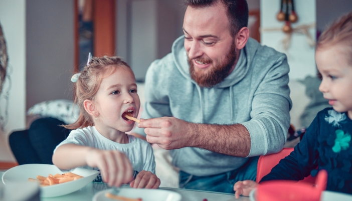 A dad feeds his young children french fries at home.