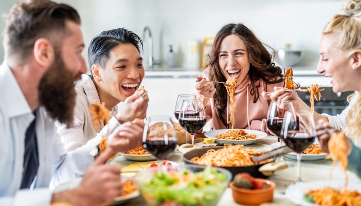 A group of friends sits down to eat a spaghetti dinner at a home.