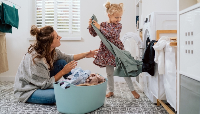 Mother and young daughter sorting a small basket of clothes in front of a front-load washer in a bright laundry room
