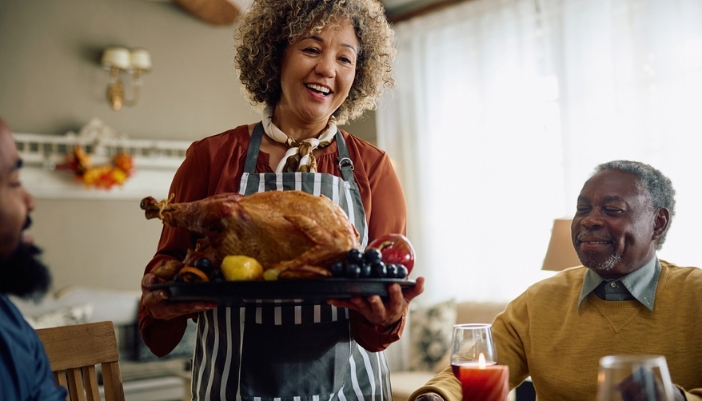 Smiling woman in an apron serving a roast turkey to family at a holiday dinner table