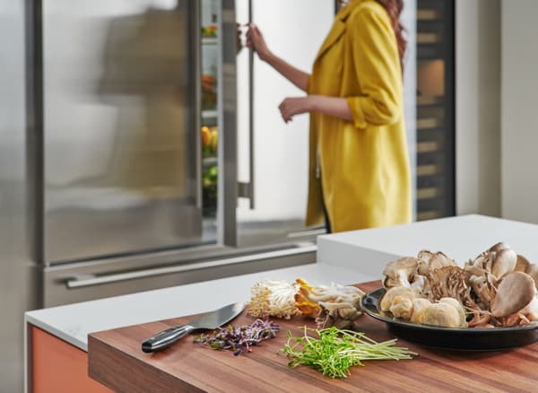 A  woman opening a Sub-Zero refrigerator in the background and a variety of mushrooms in the foreground 