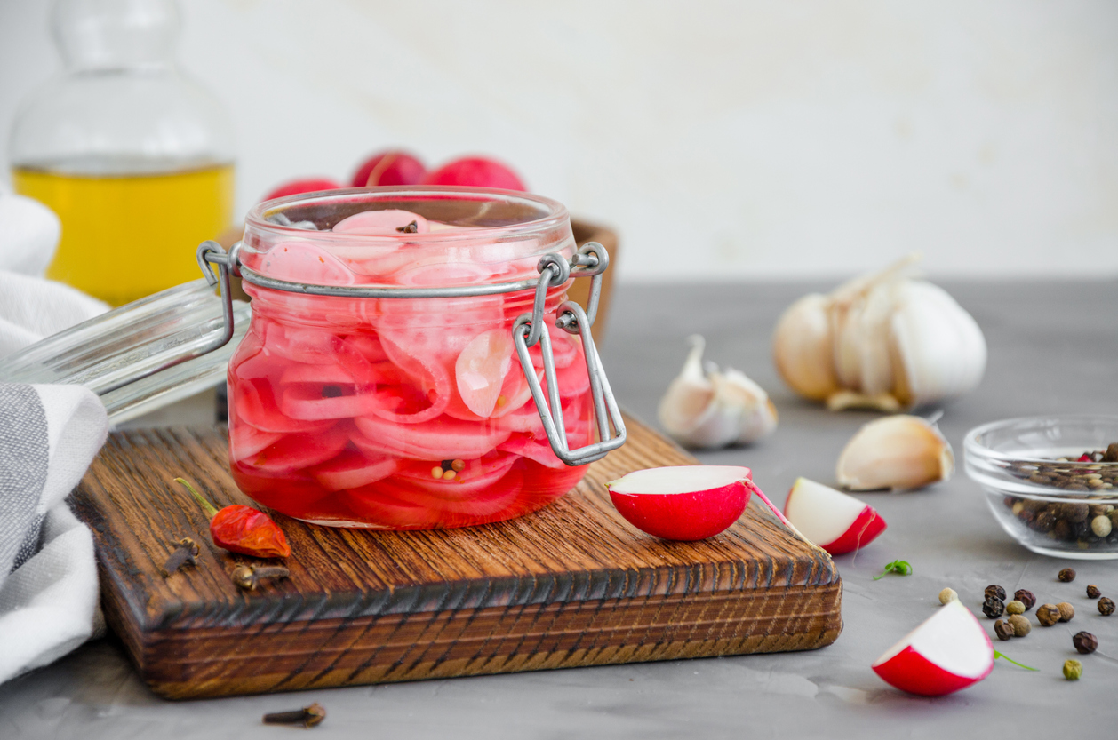 iStock Credit: Irina Taskova a small glass jar of pickled radishes sits on a cutting board near cloves and garlic