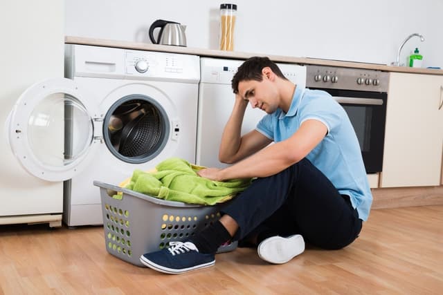 View of a man with his hand on his head sitting in front of his washer and dryer with a load of laundry