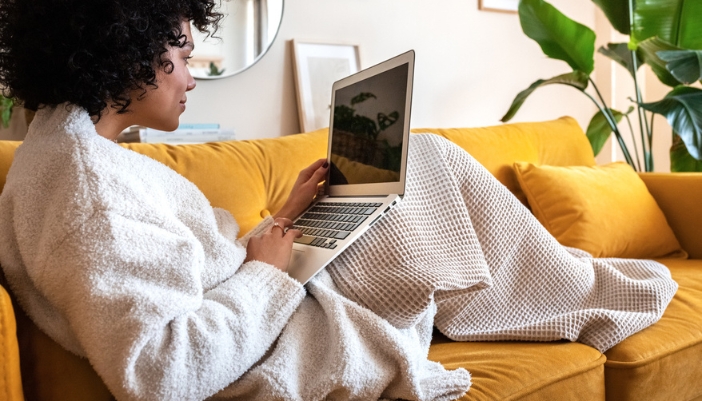 A woman sits on her bold yellow couch working on a computer.
