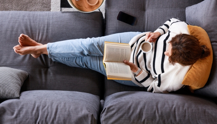 A young woman stretches out on a couch and reads her book.