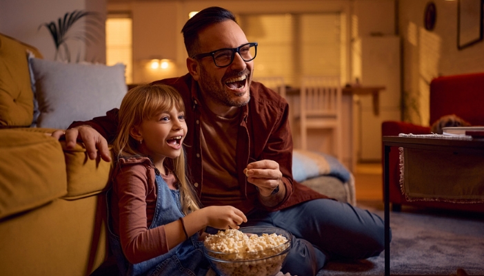 A father and daughter sit on the floor in front of their couch to watch a movie.