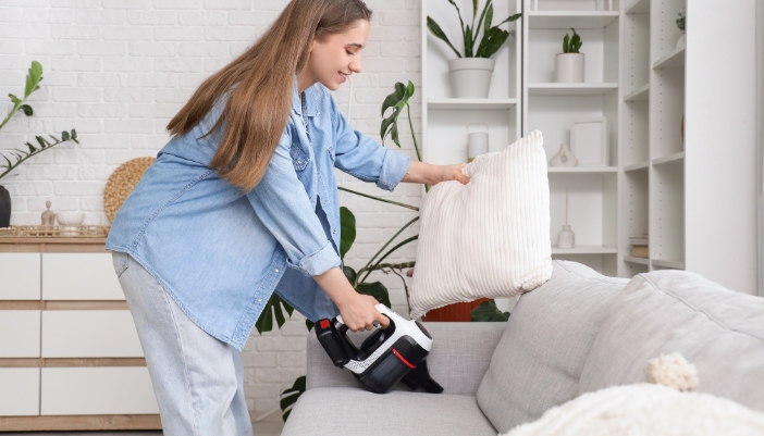 A young woman vacuums between the cushions on her couch.