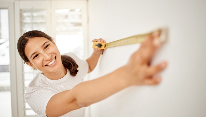 A smiling woman holds a measuring tape up to the wall.