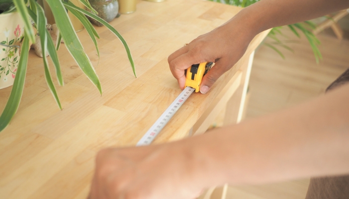 A pair of hands uses a measuring tape to measure the top of a media console.