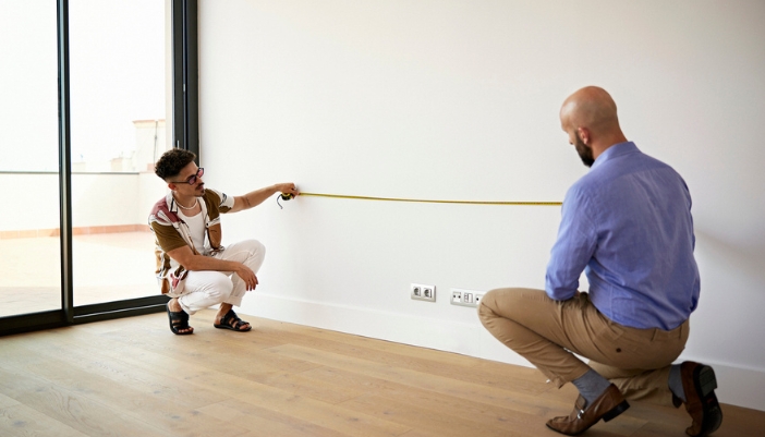 A pair of men measure against the wall in their home.