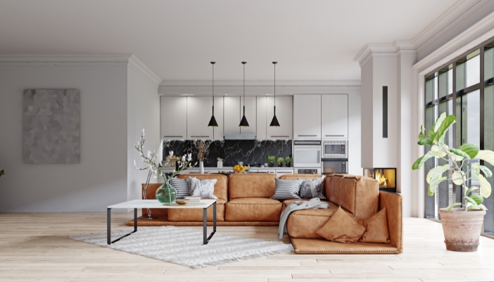 An L-shaped brown couch sit sin a naturally lit room with a kitchen behind it.