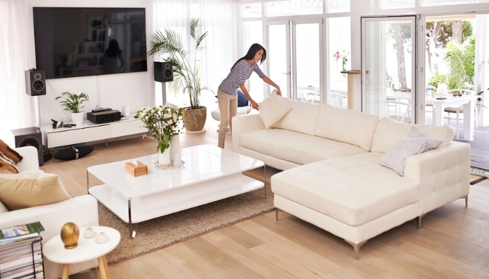 Woman adjusting a pillow on a white sectional sofa in a bright living room with floor-to-ceiling windows and a white coffee table