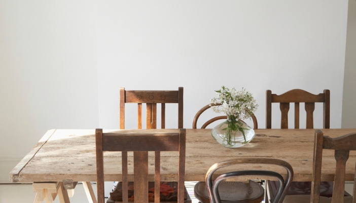 Rustic light wood dining table surrounded by mismatched vintage wooden chairs and a small vase of white flowers