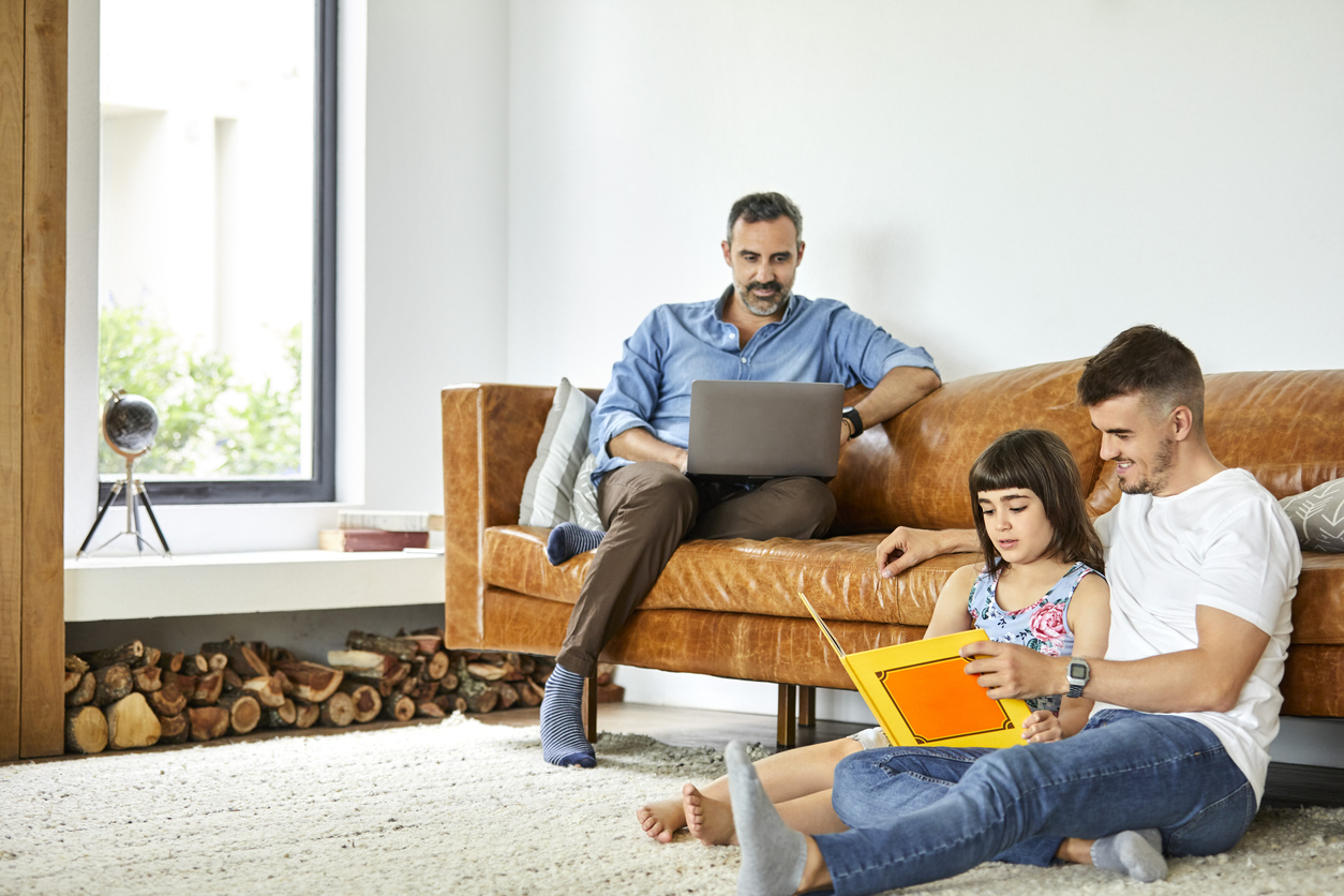 two dads and their daughter relax in a living room on a camel-colored mid-century modern couch