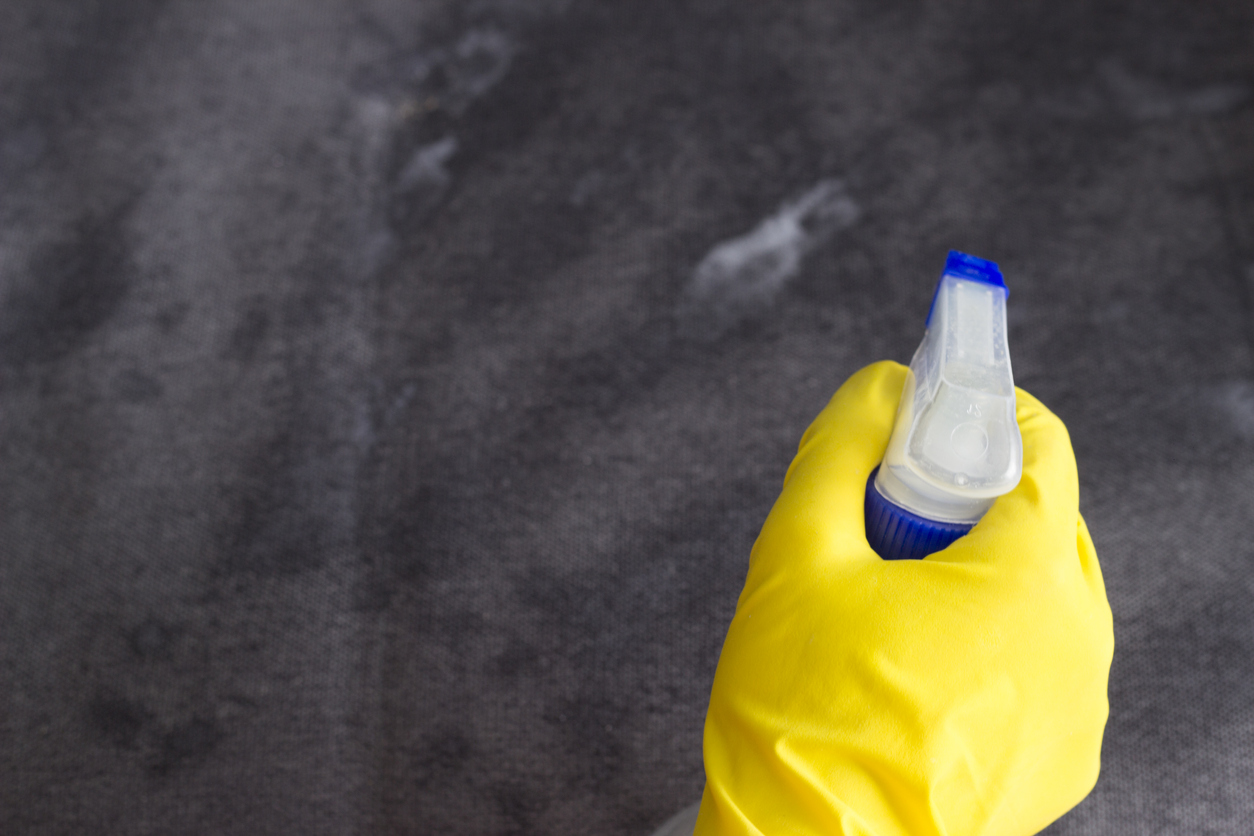 person wearing yellow dish glove sprays cleaner onto gray furniture