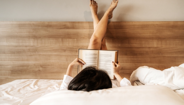 A woman lays with her feet in the air and reads in bed.