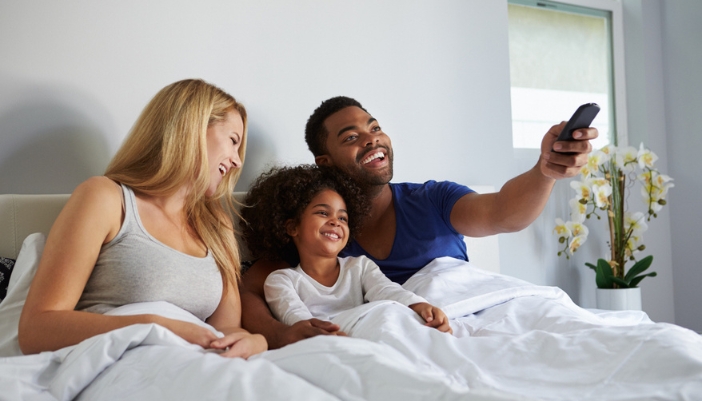 A family lays in bed happily watching TV together.