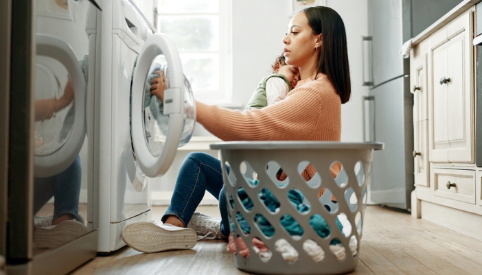 A woman holding a sleeping child empties her laundry machine at home.