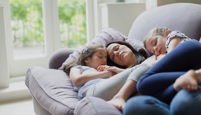 A mom naps on a couch with two daughters laying beside her and on top of her.