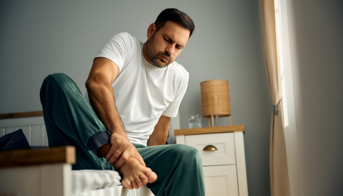 A man sits on the edge of his bed and holds his leg as if in pain.