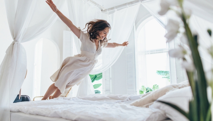 A woman jumps onto a white linen bed. 