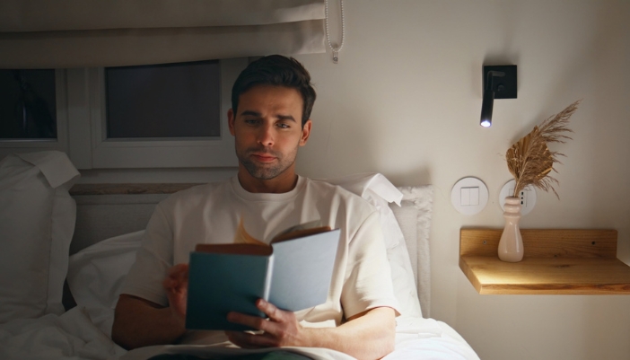 Man reading a book in bed at night under the warm glow of a wall-mounted reading light