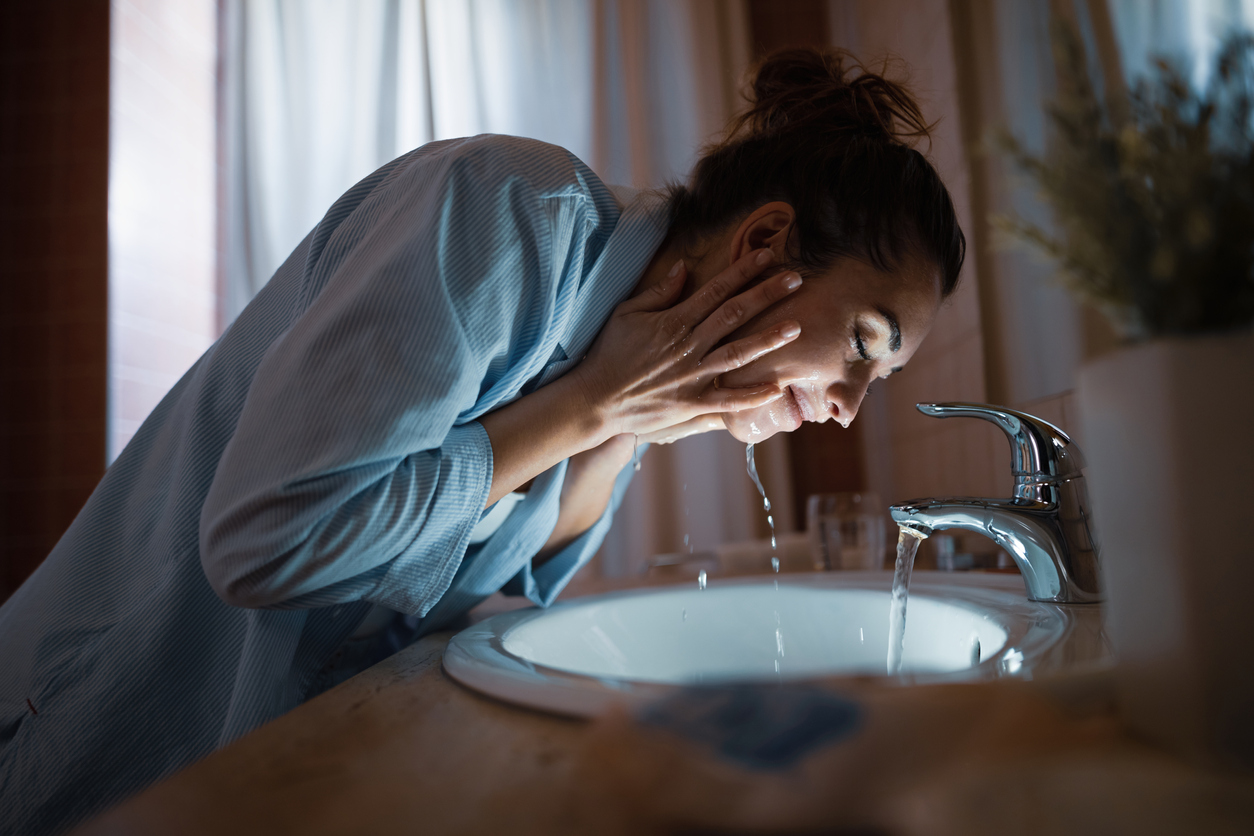 iStock Credit: Drazen Zigic young smiling woman washes her face over bathroom sink at night