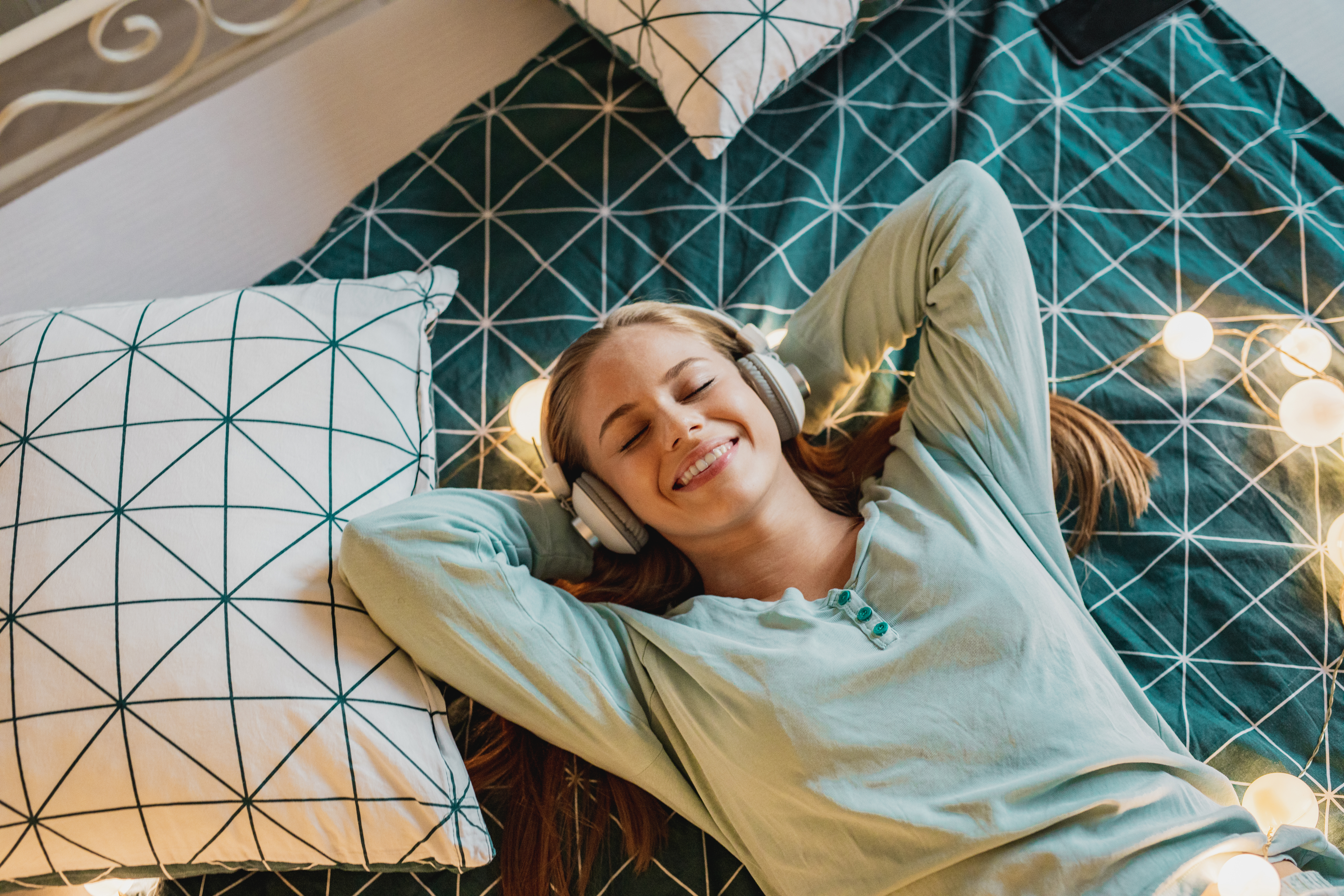 iStock Credit: Milko Young woman lying on teal and white bedding listens to music on headphones