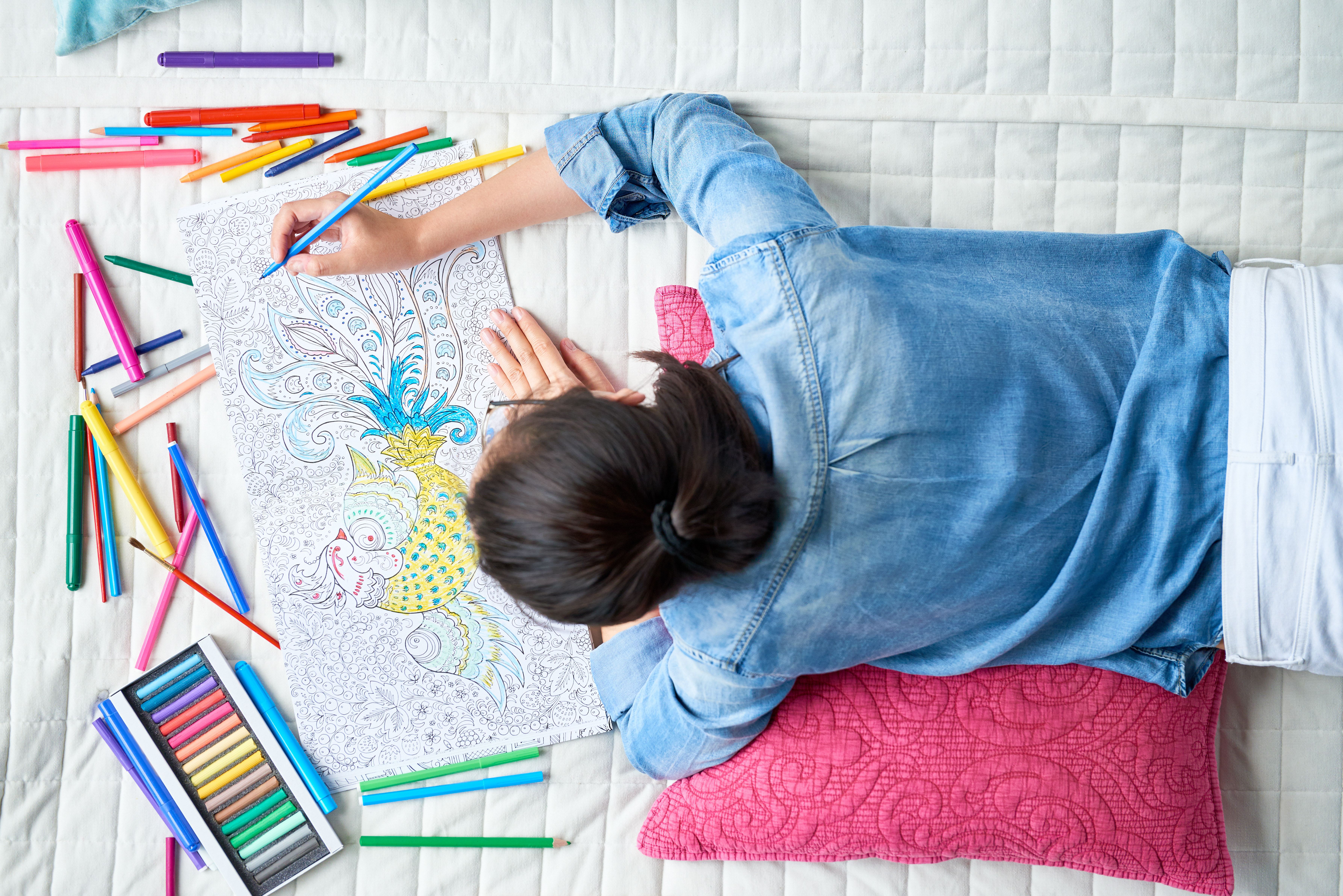 iStock Credit: mediaphotos Young woman in jean jacket colors in an adult coloring book on a bed