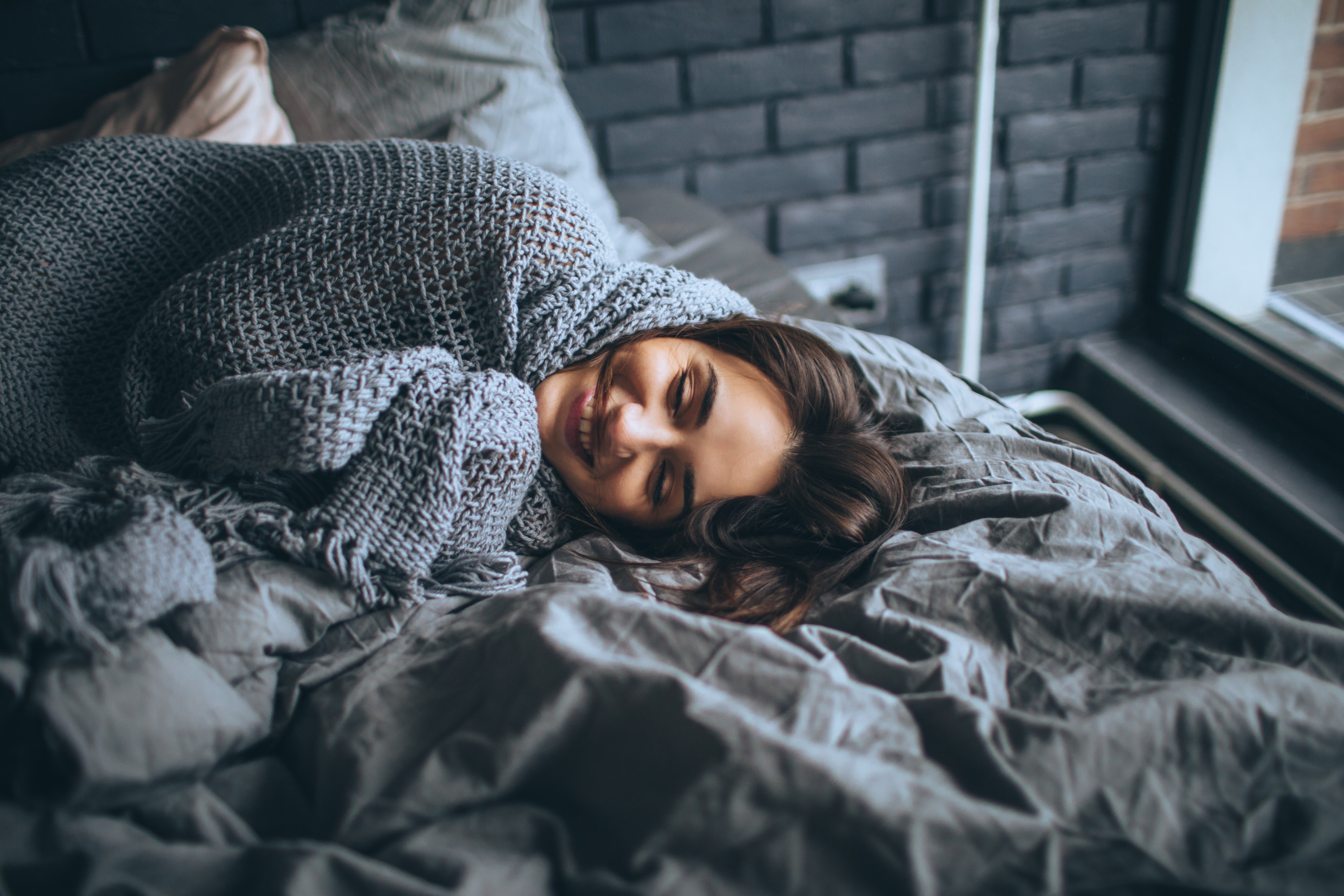 iStock Credit: AleksandarNakic young woman smiles as she wraps herself in gray knitted throw