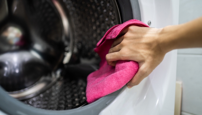 A hand uses a pink towel to clean the inside of a washing machine drum.