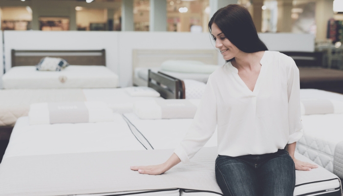 A woman sits on a mattress in a showroom.