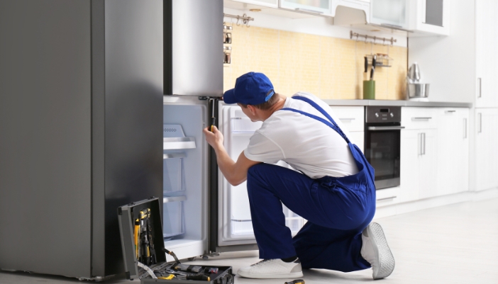 A technician in blue overalls reaches into a freezer to fix it.
