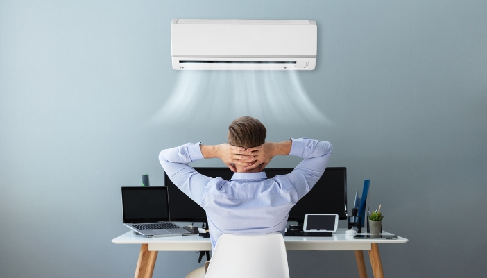 A man leans back at his desk at home under a functioning wall air conditioning unit.