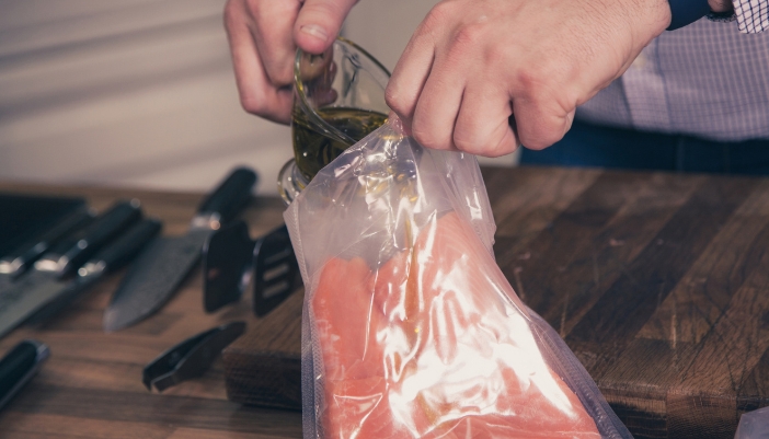 Home cook pouring olive oil into a bag with a salmon fillet on a wooden cutting board, preparing it for air sous-vide