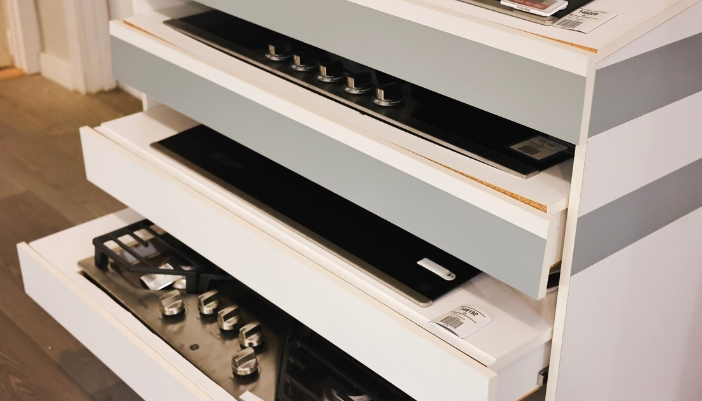 A set of white drawers in an appliance showroom with a variety of cooktops inside.
