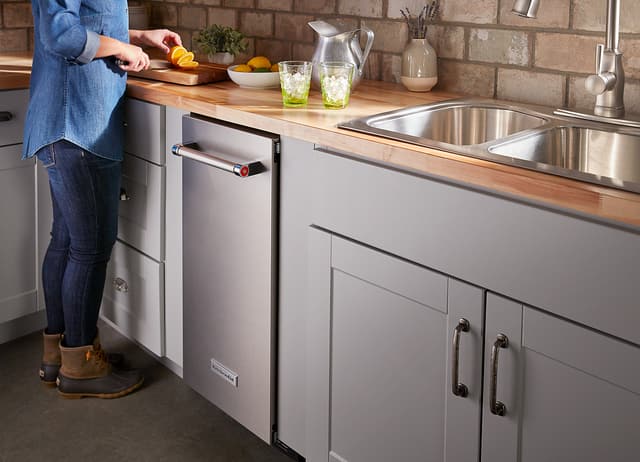 Credit: KitchenAid View of a woman cooking on a butcher block counter next to her stainless steel KitchenAid dishwasher