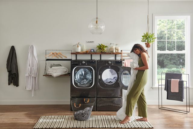 Credit: LG Front view of a modern laundry room with an LG washer and dryer and a woman standing and holding her clean, dry, sheets.