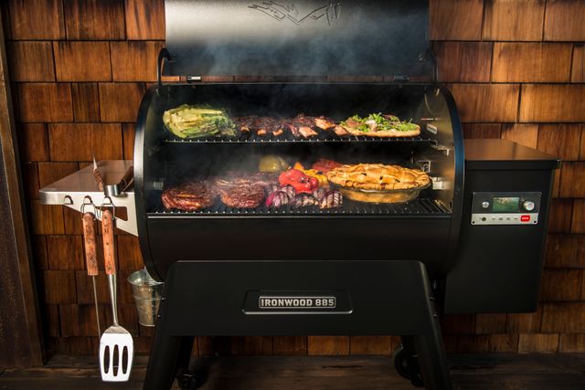 Credit: Traeger Front view of a Traeger Ironwood grill in a modern backyard, opened with meats and veggies grilling on top.