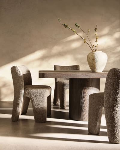 Dining set with sculptural, curved chairs in brown bouclé fabric around a dark wood table with a ribbed pedestal base, a ceramic vase on top, lit by soft sunlight.
