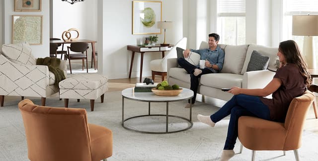 Front view of a modern living room with a sofa and three accent chairs, as a man sits on the sofa and a woman on a chair Front view of a modern living room with a sofa and three accent chairs, as a man sits on the sofa and a woman on a chair