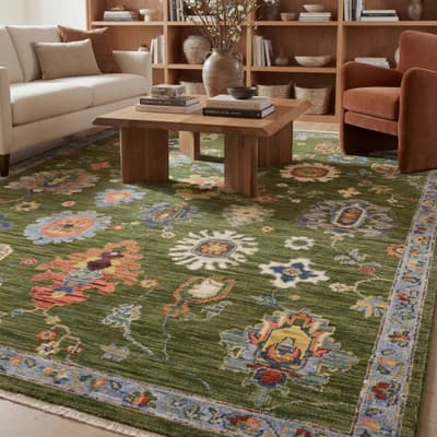 Green area rug with an oversized floral pattern in red, orange, and cream, under a live-edge wood coffee table stacked with books, near a cream sofa and rust accent chair.